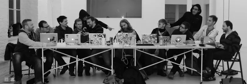 A group of men sitting at a long table with their laptops, some full of stickers. The picture depicts a situation that looks like the last supper, or more likely: 'the last coding session'.