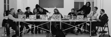 A group of men sitting at a long table with their laptops, some full of stickers. The picture depicts a situation that looks like the last supper, or more likely: 'the last coding session'.