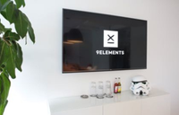 There is a TV showing the 9elements logo hanging over a white tv-board with a Stormtrooper helmet, drinks and glasses on it. A plant protrudes a little from the left into the picture.
