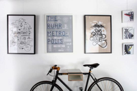 A wall on which hang three larger and three smaller illustrated pictures. Below is a wooden bracket on the wall on which is hung a bicycle