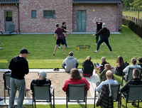 Four men are playing spikeball in the background, in the foreground many people are sitting on a green lawn and chairs watching.